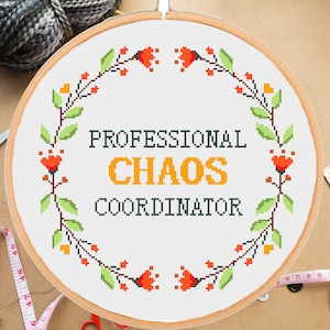 Puede incluir: Un aro de bordado blanco con un diseño de corona floral y el texto "PROFESSIONAL CHAOS COORDINATOR" en letras negras y naranjas.
