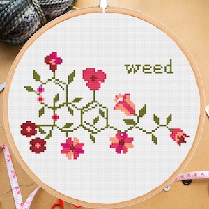 May include: A cross-stitch embroidery hoop with a floral design and the word "weed" stitched in green thread. The flowers are pink, red, and orange.