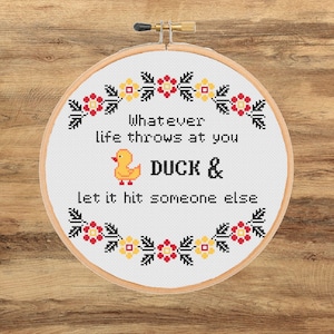 Può includere: Un telaio da ricamo a punto croce con la scritta "Whatever life throws at you DUCK & let it hit someone else." Il disegno comprende un'anatra gialla e un bordo floreale rosso, giallo e nero. Il telaio è posto su uno sfondo di legno.