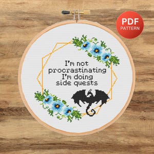 May include: Cross stitch pattern featuring a black dragon and blue flowers with the text "I'm not procrastinating I'm doing side quests".