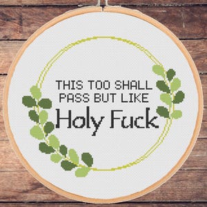 Può includere: Un telaio rotondo per punto croce bianco con il testo "THIS TOO SHALL PASS BUT LIKE Holy Fuck" in nero. Il design include un bordo floreale verde e giallo. Lo sfondo è una superficie di legno.