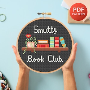 May include: A circular wooden embroidery hoop containing a cross-stitch design on a dark gray fabric. The text reads Smutty Book Club. The design features a brown shelf with a potted plant, four books, and a patterned spine.