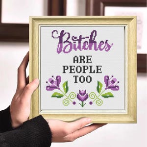 May include: Cross-stitch artwork in a light wood frame. The text reads "Bitches ARE PEOPLE TOO" in purple and black. Floral accents in purple and green surround the text, creating a decorative design.