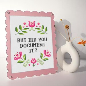 May include: A pink framed cross-stitch with the text "BUT DID YOU DOCUMENT IT?" surrounded by floral embroidery. A white vase with a circular opening holds dried flowers. The frame has a scalloped edge.