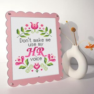 以下が含まれることがあります： 「Don't make me use my HR voice」の文字と花柄が刺繍されたピンク色の額縁。ユニークな形の白い花瓶にはドライフラワーが飾られ、装飾的なアクセントを添えています。