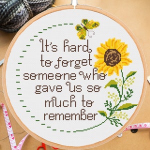 May include: A white embroidery hoop with a cross-stitch design featuring a sunflower, butterfly, and the text "It's hard to forget someone who gave us so much to remember".