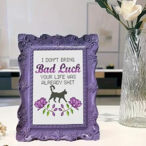 May include: A rectangular purple ornate frame containing a cross-stitch piece with the text 'I DON'T BRING Bad Luck YOUR LIFE WAS ALREADY SHIT' in black and purple lettering, featuring a black cat silhouette and two purple roses with green leaves.