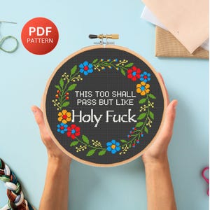 Puede incluir: Un bastidor de bordado de punto de cruz con el texto "THIS TOO SHALL PASS BUT LIKE Holy Fuck" rodeado por una corona floral. El diseño presenta flores rojas, azules y amarillas sobre un fondo negro. Un círculo rojo con "PDF PATTERN" está en la parte superior izquierda.