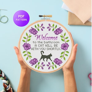 Può includere: Un telaio da ricamo a punto croce con la scritta "Benvenuti in bagno, un gatto sarà con voi a breve". Il disegno comprende una sagoma di gatto nero, fiori viola e foglie verdi. Un cerchio viola con "PDF PATTERN" è in alto a sinistra.