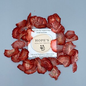 May include: Dried strawberry slices arranged around a card with the text "HOPE'S HEALTHY TREATS". The card also includes a small illustration of a hamster. The strawberries are a deep red color and are thinly sliced.