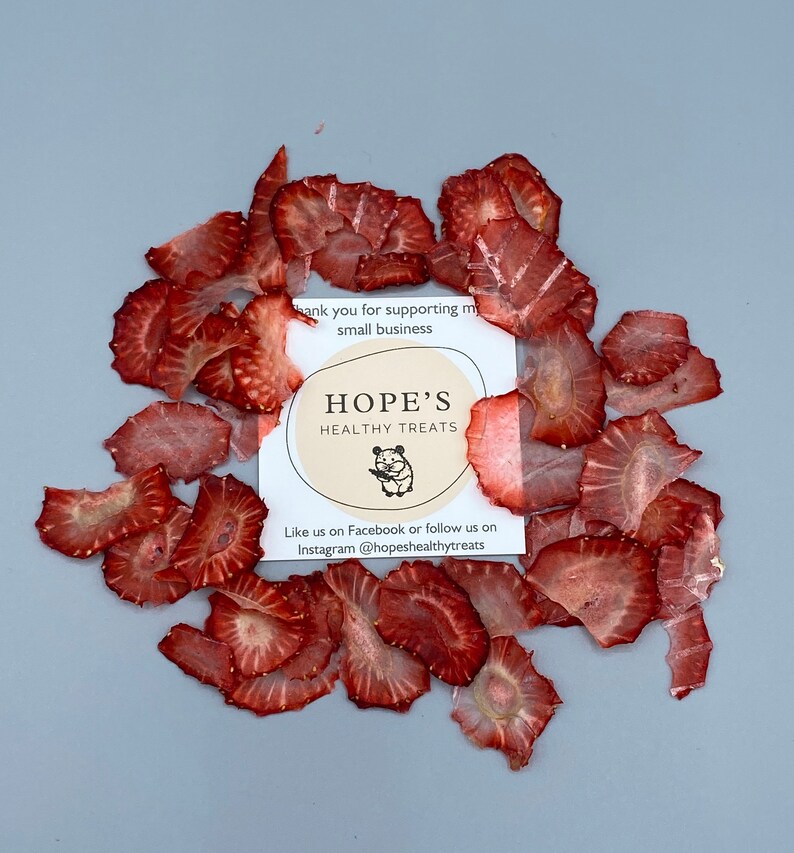 May include: Dried strawberry slices arranged around a card with the text "HOPE'S HEALTHY TREATS". The card also includes a small illustration of a hamster. The strawberries are a deep red color and are thinly sliced.
