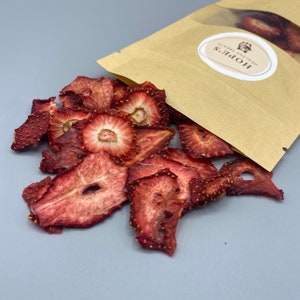 May include: Dried strawberry slices spilling from a brown paper bag. The strawberries are a deep red color with visible seeds and a slightly translucent appearance. The bag has a white oval sticker with the text "HOPE'S HEALTHY TREATS".