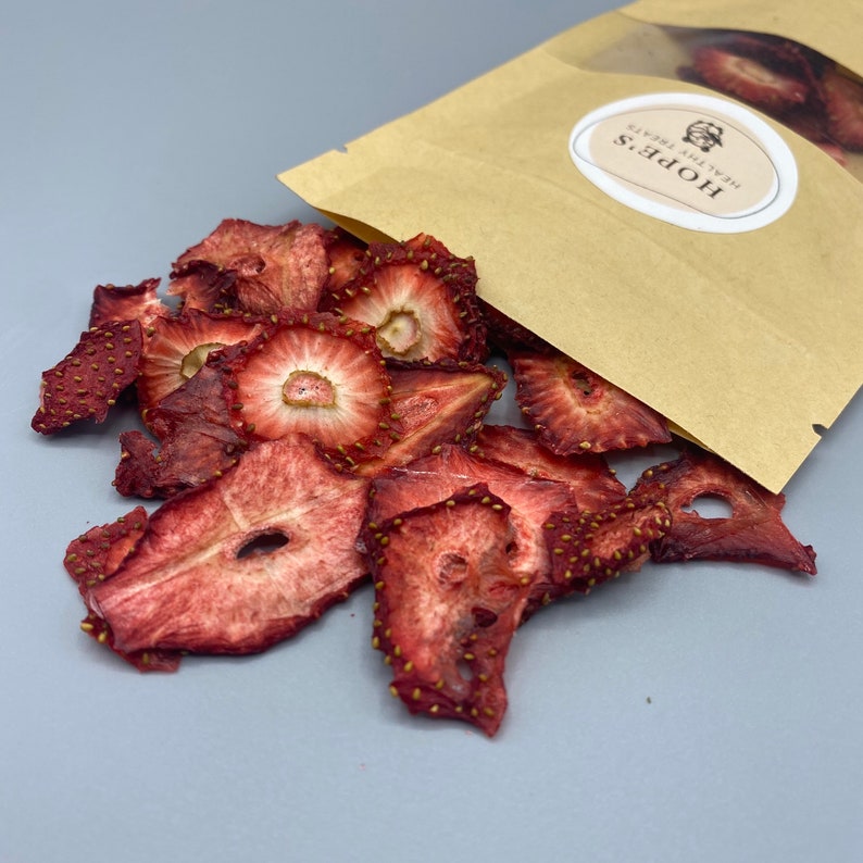 May include: Dried strawberry slices spilling from a brown paper bag. The strawberries are a deep red color with visible seeds and a slightly translucent appearance. The bag has a white oval sticker with the text "HOPE'S HEALTHY TREATS".