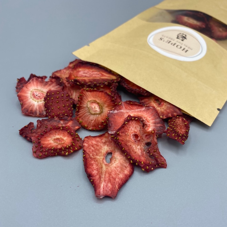 May include: Dried strawberry slices spilling from a brown paper bag. The strawberries are a deep red color with visible seeds around the edges. The bag has a white oval label with the text "HOPE'S HEALTHY TREATS".