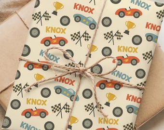 May include: Gift-wrapped present with a beige paper featuring a pattern of red, blue, and yellow race cars, checkered flags, and trophies. The word "KNOX" is printed throughout the design. The gift is tied with twine and sits on a brown paper.