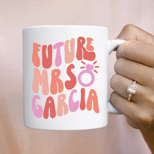 May include: White ceramic mug with the text "FUTURE MRS GARCIA" in shades of pink and red. A pink ring with a heart detail is also featured. The mug is held by a person wearing a ring.