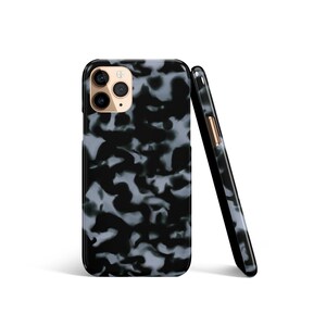 Peut inclure: Deux coques de téléphone à motifs noirs et gris. L'une est droite, montrant l'appareil photo et le flash. L'autre est appuyée contre elle, révélant une vue latérale avec un bouton doré. Les coques ont un motif marbré.