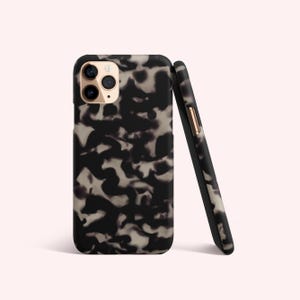 Puede incluir: Dos fundas de teléfono con estampado negro y beige. Una funda está en posición vertical, mostrando las lentes de la cámara y el flash. La otra funda está apoyada contra la primera, mostrando el lateral con un botón dorado. Las fundas tienen un diseño de carey.