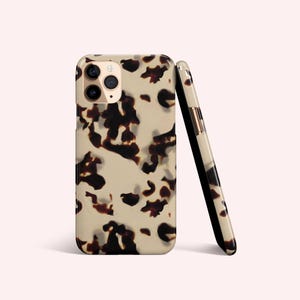 May include: Two phone cases with a tortoiseshell pattern. The cases are beige with dark brown spots. One case is upright, showing the camera lenses, while the other is leaning against it, displaying the side.