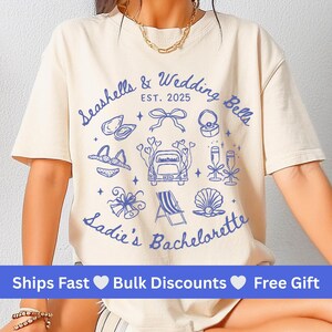 Peut inclure: Un t-shirt crème avec un motif graphique bleu représentant des coquillages, des cloches de mariage, une voiture avec un panneau 'Just Married' et le texte 'Seashells & Wedding Bells EST. 2025 Sadie's Bachelorette'.