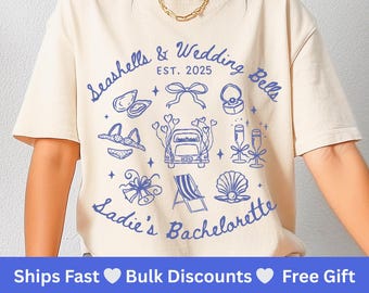 May include: A cream-colored t-shirt with a blue graphic design featuring seashells, wedding bells, a car with a 'Just Married' sign, and the text 'Seashells & Wedding Bells EST. 2025 Sadie's Bachelorette'.