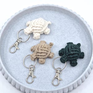 May include: Three crocheted turtle keyrings, one white, one beige, and one dark green, with silver key rings and clips.
