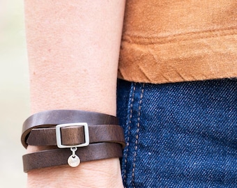 May include: A dark brown leather wrap bracelet featuring a silver-toned rectangular buckle and a small circular charm. The bracelet is worn on the wrist, paired with a plain tan short-sleeved top and dark blue denim jeans with visible pockets.