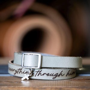 May include: A light gray leather wrap bracelet featuring a silver-toned rectangular buckle and a small silver cross charm. The leather band is engraved with the cursive text "everything through him" in a dark font, creating a layered appearance.