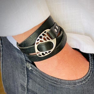 May include: A black leather wrap bracelet with a silver, openwork, oval-shaped charm. The bracelet is wrapped around a person's wrist.