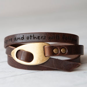 Peut inclure: Un bracelet en cuir marron à enrouler avec une fermeture en métal doré et deux rivets en métal doré. Le bracelet porte l'inscription "Inspire and others will follow" gravée.