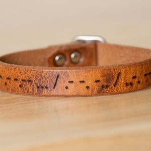 May include: A brown leather bracelet with Morse code etched into the surface. The bracelet has a buckle closure and is approximately 0.75 inches wide. The leather has a distressed appearance, adding to its rustic charm.