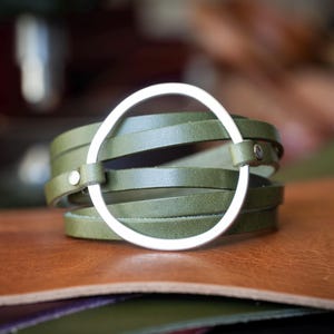 May include: Olive green leather wrap bracelet with a silver metal circle accent. The bracelet is made of multiple leather straps that wrap around the wrist. The silver circle is attached to the leather straps with small silver rivets. The bracelet is displayed on a brown leather surface.