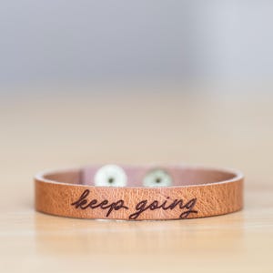 May include: A tan leather bracelet with the words "keep going" engraved on the front. The bracelet has two silver-coloured snap closures. The bracelet is approximately 2 cm wide.