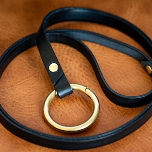 May include: A black leather lanyard with a gold metal ring. The lanyard is coiled on a brown leather surface.