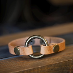 May include: A light brown leather bracelet featuring a large, circular, antique brass-coloured ring. The bracelet has brass-coloured button closures. The item is displayed on a wooden surface. The bracelet is approximately 20 cm in length.