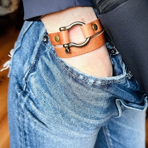 Peut inclure: Une ceinture en cuir marron avec une boucle en métal. La ceinture est portée autour de la taille et rentrée dans la poche avant d'un jean bleu.
