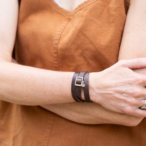 May include: A dark brown leather wrap bracelet featuring a silver-toned rectangular buckle and a small circular charm. The bracelet is worn around the wrist, displaying a smooth finish and a multi-layered design that secures with a metal clasp.