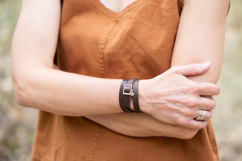 May include: A dark brown leather wrap bracelet featuring a silver-toned rectangular buckle and a small circular charm. The bracelet is worn around the wrist, displaying a smooth finish and a multi-layered design that secures with a metal clasp.