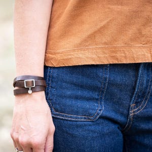 May include: A dark brown leather wrap bracelet featuring a silver-toned rectangular buckle and a small circular charm. The bracelet is worn on the wrist, paired with a plain tan short-sleeved top and dark blue denim jeans with visible pockets.