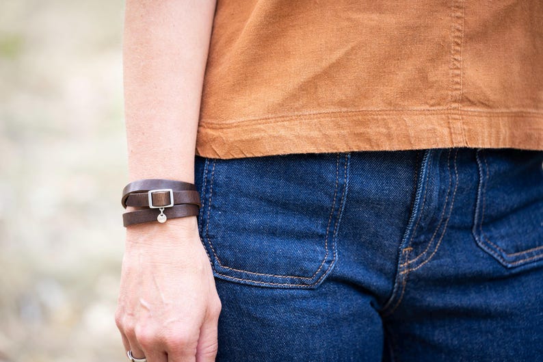 May include: A dark brown leather wrap bracelet featuring a silver-toned rectangular buckle and a small circular charm. The bracelet is worn on the wrist, paired with a plain tan short-sleeved top and dark blue denim jeans with visible pockets.