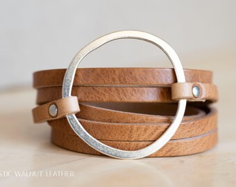Könnte beinhalten: Wickelarmband mit mehreren braunen Lederriemen und einem silberfarbenen Metallring. Die Lederriemen sind mit kleinen, hellbraunen Lederschlaufen und silberfarbenen Metallknöpfen befestigt. Ein neutrales Accessoire.