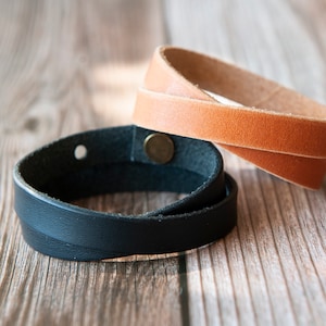 May include: Two leather bracelets, one black and one brown, are shown on a wooden surface. The black bracelet has a small metal button closure.
