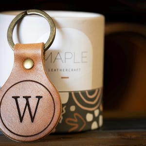 Op de afbeelding: Een beige leren sleutelhanger met een cirkelvormig ontwerp en de letter W in het zwart in het midden gedrukt. De sleutelhanger is met een messingkleurige klinknagel aan een metalen ring bevestigd. Het leer heeft een gladde afwerking.