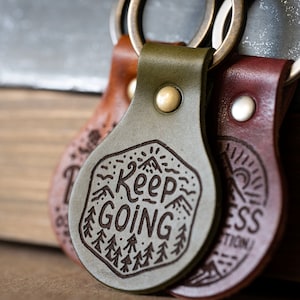 Könnte beinhalten: Drei Leder-Schlüsselanhänger mit Metallringen. Die Schlüsselanhänger sind grün, braun und dunkelbraun. Der grüne Schlüsselanhänger hat den Text "Keep Going" eingraviert. Der braune Schlüsselanhänger hat den Text "Happiness" eingraviert.