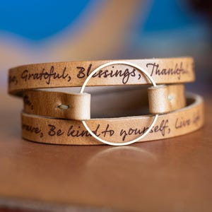 May include: A stack of three light brown leather wrap bracelets. Each bracelet is engraved with inspirational words in a handwritten font. A gold-colored metal ring connects the ends of the bracelets.