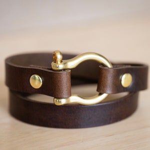 May include: A brown leather bracelet with a gold metal clasp. The bracelet is double-wrapped and has two gold metal rivets.