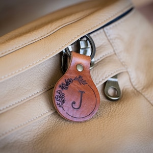 K&ouml;nnte beinhalten: Eine beigefarbene Bauchtasche aus Leder mit einem runden Rei&szlig;verschlussanh&auml;nger aus mittelbraunem Leder. Der Anh&auml;nger ist mit dem Buchstaben J in einem Kreis graviert, verziert mit einem zarten Blumenrankenmuster und einem Metallniet.