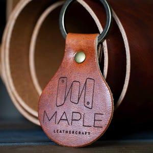 May include: A brown leather keychain with a metal ring. The keychain has the engraved text "MAPLE LEATHERCRAFT" and a logo. The background shows rolled leather.