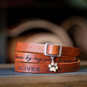 May include: A tan leather wrap bracelet featuring a silver-toned buckle and a dangling silver paw print charm. The leather is engraved with the text "Once by my side, forever" and the name "OLIVER" followed by the dates "2003 - 20015".