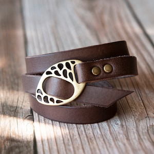 May include: A brown leather wrap bracelet with a gold-colored, oval-shaped buckle. The buckle has a decorative cutout design. The bracelet is wrapped around itself and secured with two small, gold-colored rivets.
